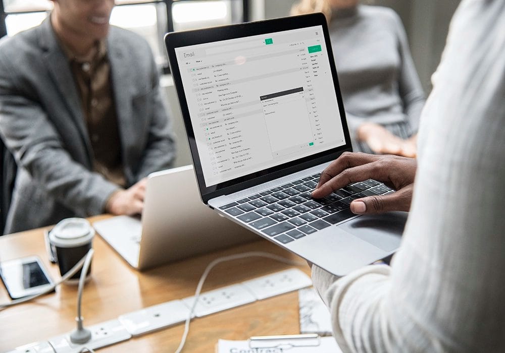 A person is using a laptop to review marketing email tips while sitting at a table with other people.