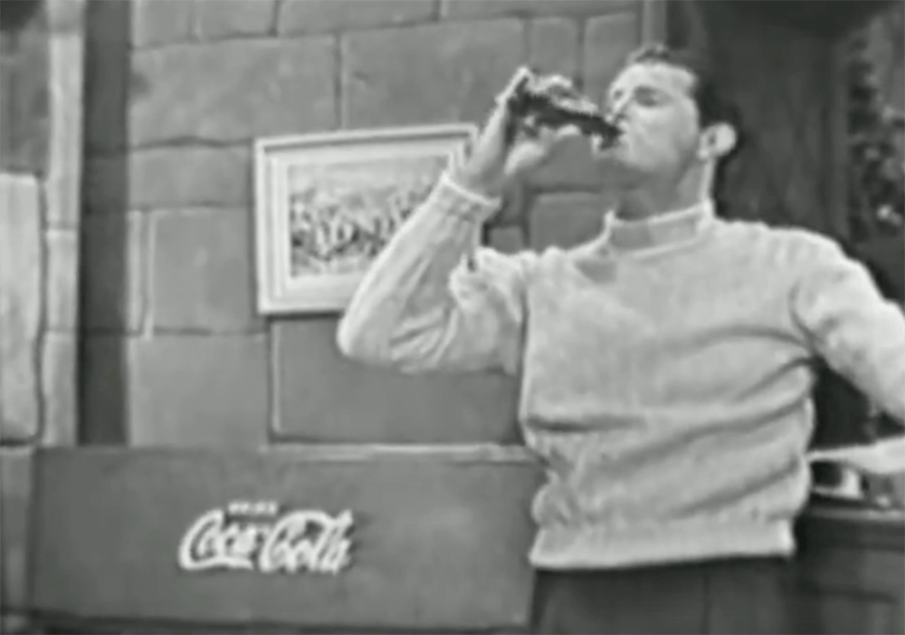 A man in a sweater drinks from a Coca-Cola bottle in a room with a stone wall and framed artwork, as part of the Coca-Cola campaigns.