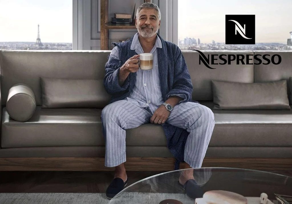 George Clooney in pajamas holding a coffee mug in a modern living room with the Eiffel Tower visible through the window, featuring Nespresso's most successful celebrity endorsement logo in the corner.