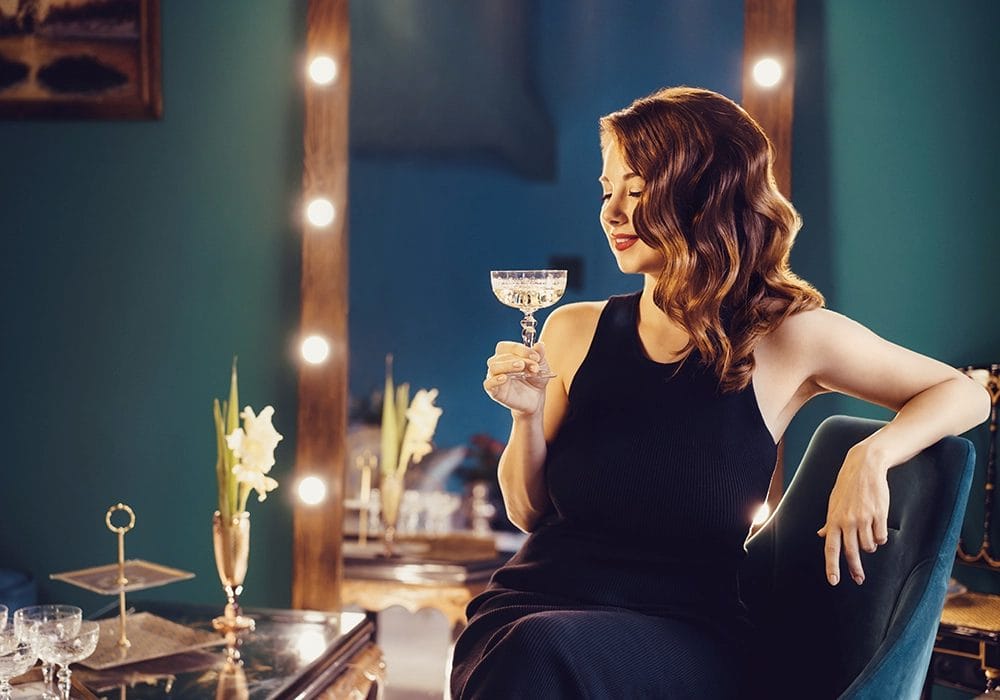 actress in a black dress sitting in a lounge, smiling and holding a cocktail glass, with illuminated vanity mirrors in the background for an TV advertisement.