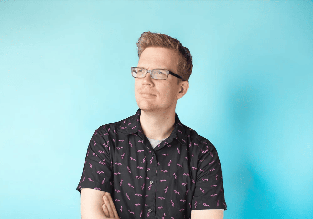 Hank Green wearing a black shirt adorned with pink flamingos, influenced by the power of influencers, looking to the side against a blue background.