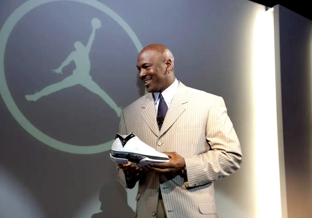Michael Jordan in a pinstripe suit smiling as he examines a basketball shoe, standing in front of the large Nike logo featuring a silhouette of a basketball player.