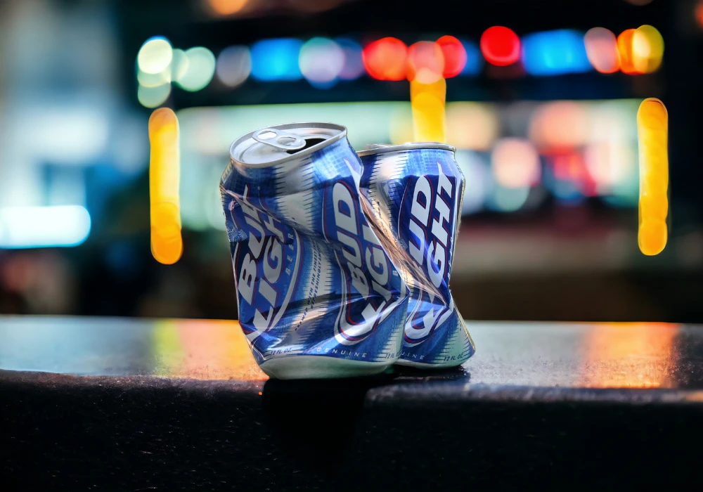 Two crushed Bud Light beer cans sit on a surface with a colorful bokeh background of blurred lights, reminiscent of the vibrant scenes seen in Budweiser's celebrity endorsements.