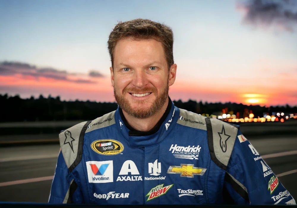 Dale Earnhardt Jr. in a blue racing suit with various sponsor logos, hinting at NASCAR celebrity endorsements, stands smiling with a sunset and track in the background.