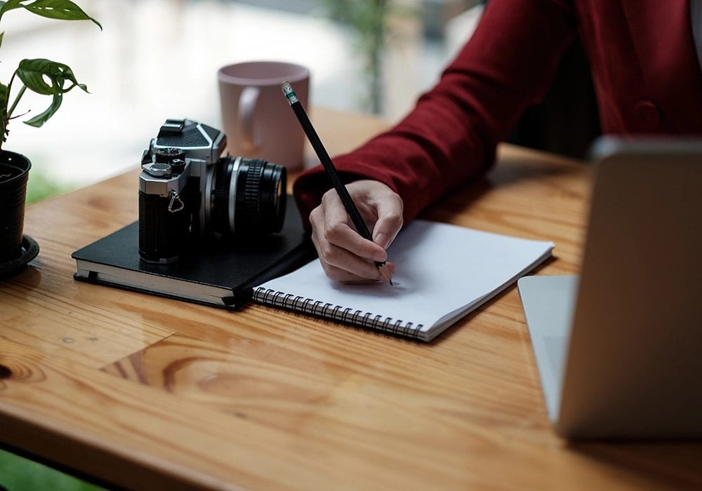 A person writes in a notebook at a wooden table, with a camera, plant, coffee mug, and laptop also on the table.