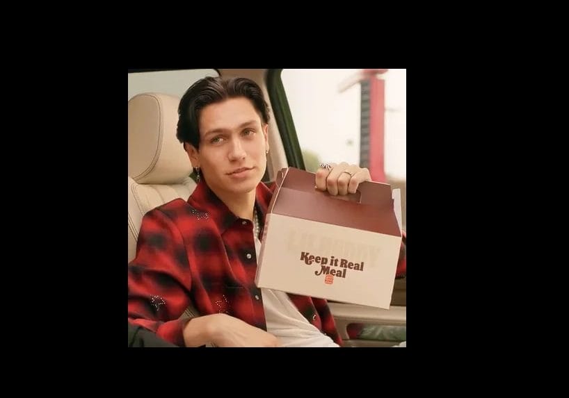 Lil Hudd sitting in a car holds up a "Keep it Real Meal" box while slightly smiling, showcasing the latest in celebrity food endorsements.