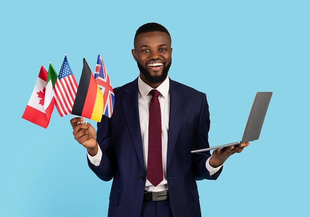 A man in a suit holds a laptop in one hand and small international flags, including Canadian, American, German, and British flags, in the other. The solid blue background highlights his focus on international digital marketing.