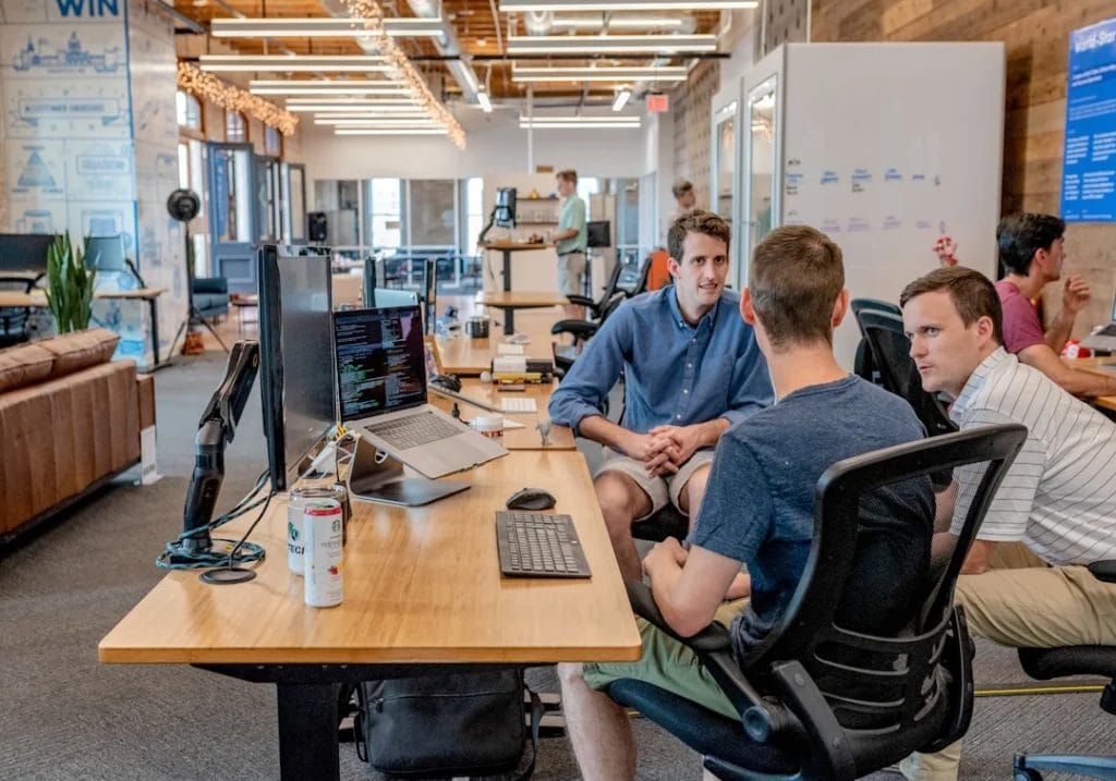 Three people sit at a wooden desk discussing work; two computer monitors and a soda can are on the desk. Other individuals work in the background within a modern office environment, brainstorming strategies for their digital marketing side hustle.