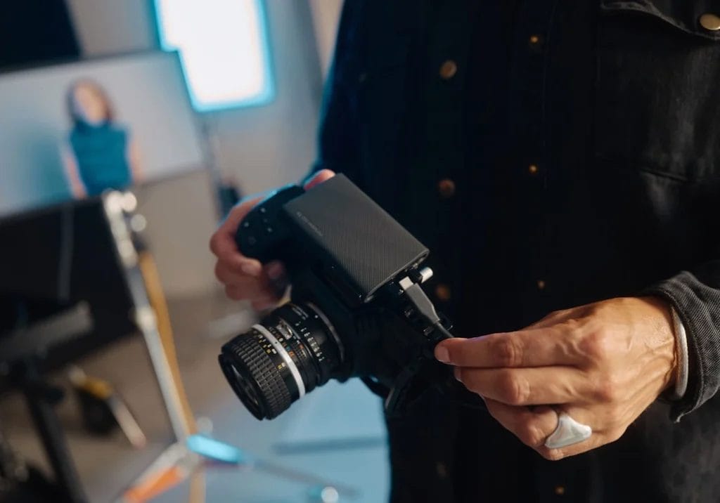 Person adjusting settings on a professional camera in a photography studio setup, likely for their digital marketing side hustle. A model is blurred in the background.