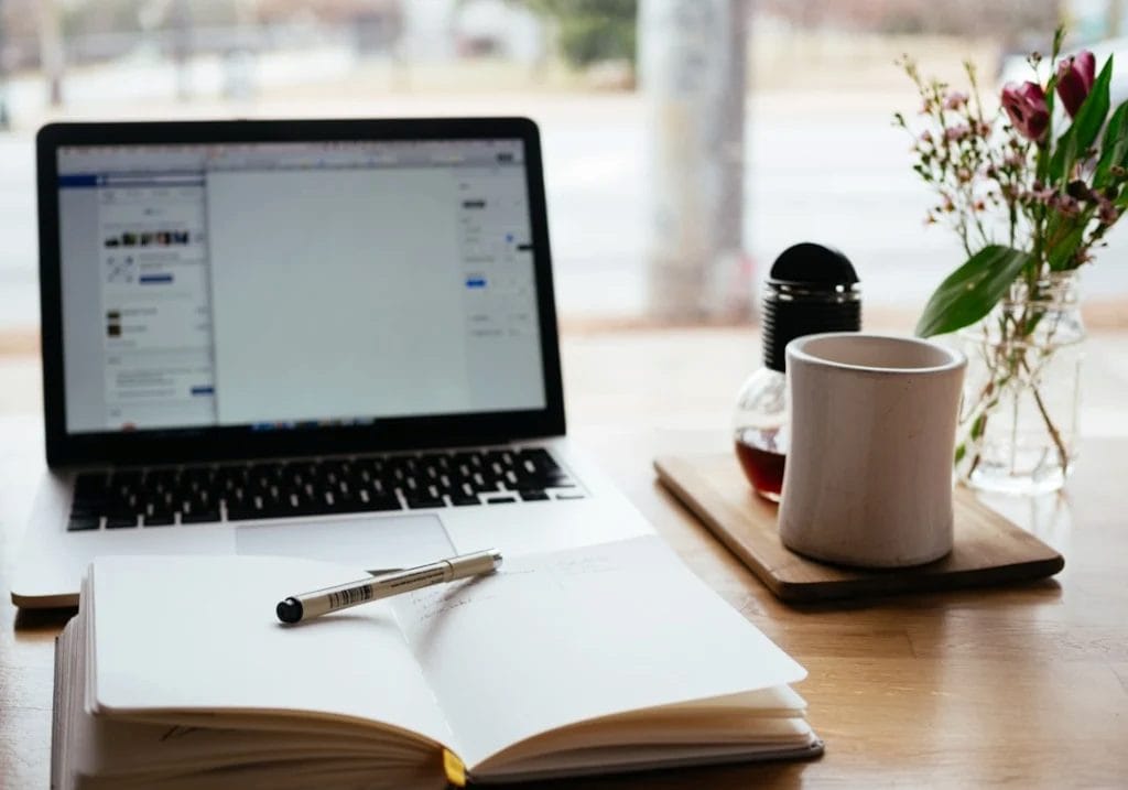 A laptop displaying a digital marketing side hustle webpage, an open notebook with a pen, a coffee cup on a wooden tray, and a vase with flowers are arranged neatly on the wooden desk.