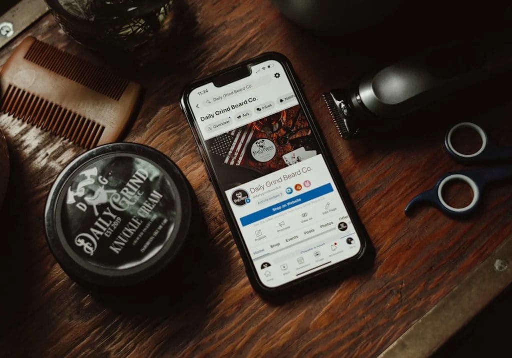 A smartphone with a beard care company's social media page displayed, surrounded by grooming tools including a container of beard balm, a comb, a trimmer, and scissors on a wooden surface—perfect for anyone exploring digital marketing as their side hustle.