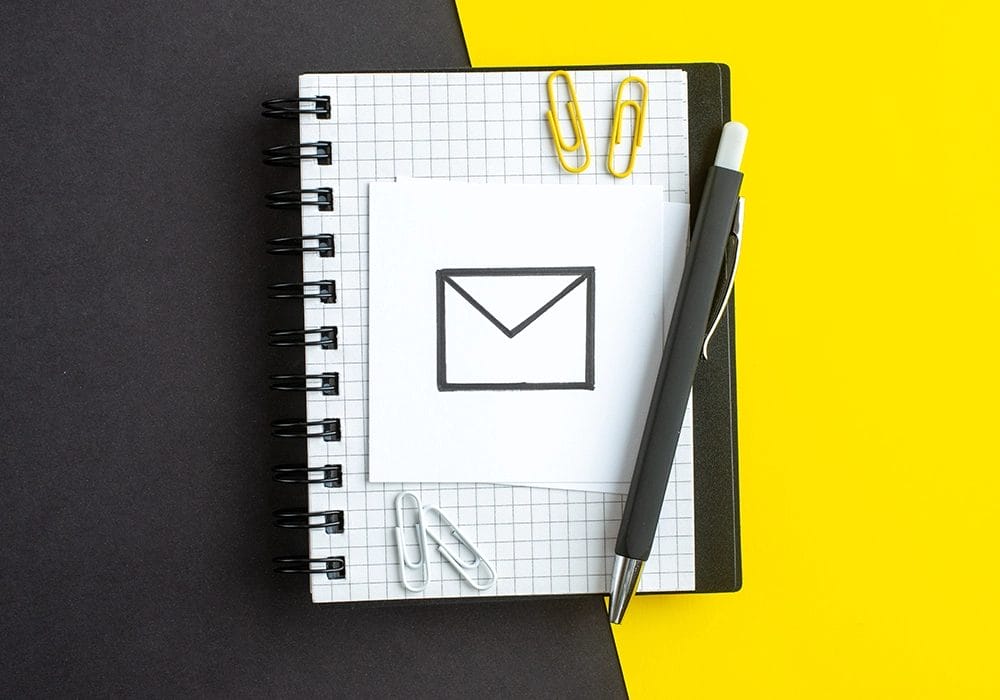 A spiral notebook lies open with a paper displaying an envelope symbol, suggesting the power of Email Marketing. A black pen rests on the notebook, with several paper clips scattered around against a split black and yellow background.