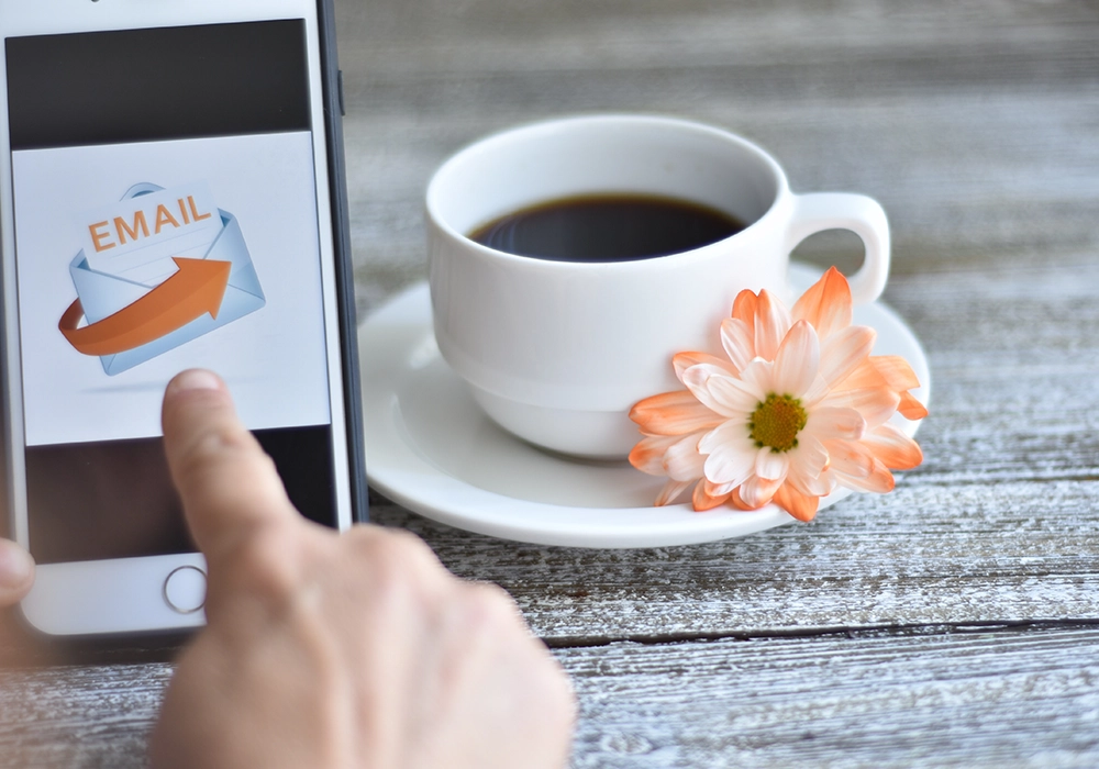 A person uses a smartphone displaying an email icon next to a white cup of coffee with a peach and white flower on the saucer, placed on a wooden surface, emphasizing how Email Marketing Works seamlessly even in everyday moments.