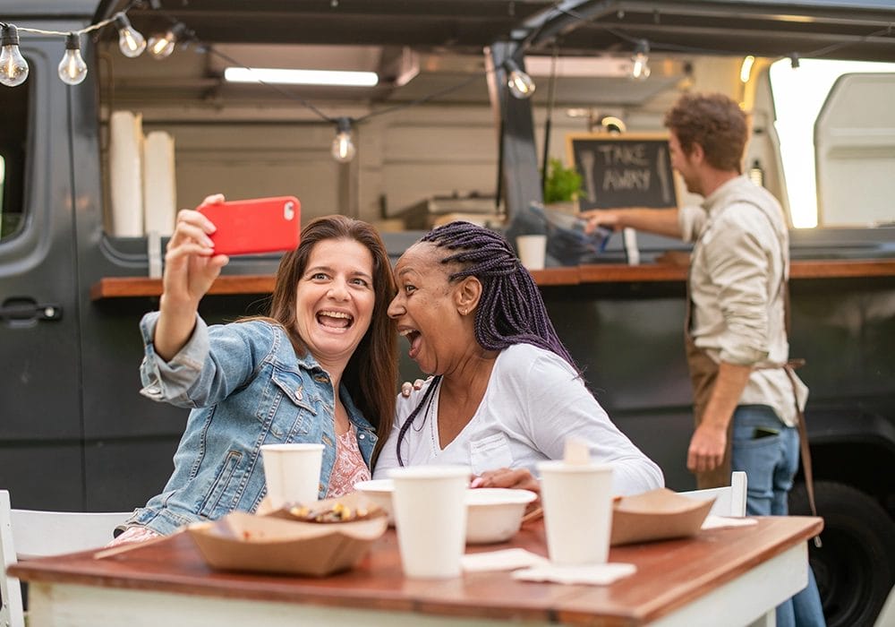 Two women are sitting at a table with food and drinks, taking a selfie and smiling, providing perfect social proof of their enjoyable outing. Meanwhile, a man is busy working at a food truck in the background.