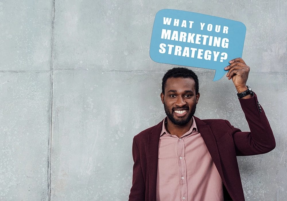 A man stands against a concrete wall, holding a blue speech bubble sign that reads, "What is your customized digital marketing strategy?