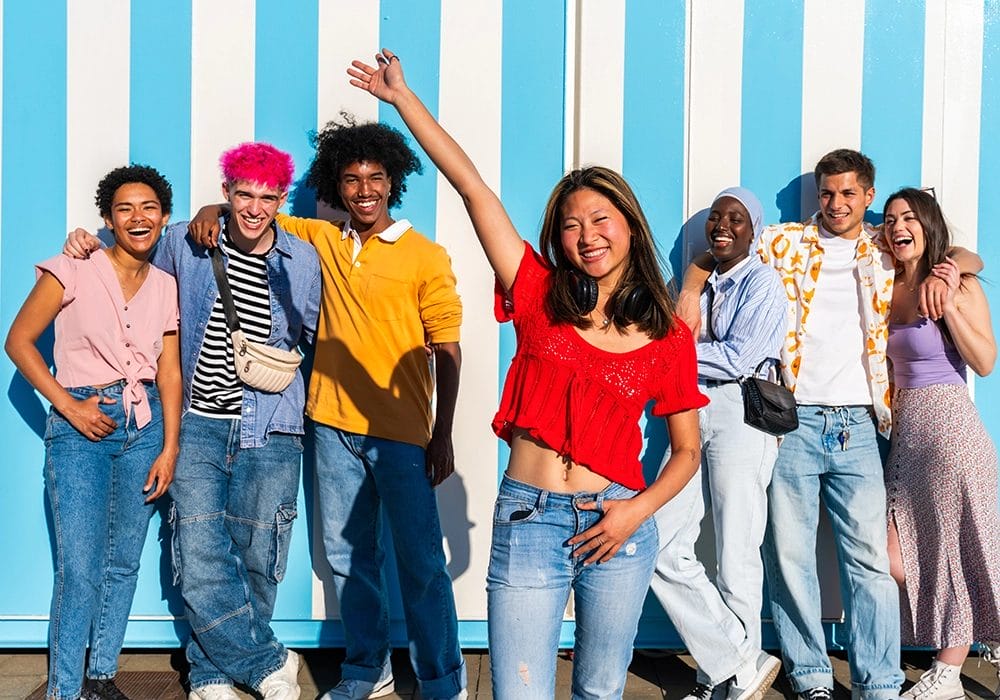 A diverse group of seven young adults smiling and posing for a photo against a blue and white striped background showcases social proof. One person in a red top stands in the front with a raised arm, emphasizing their mean but charismatic presence.