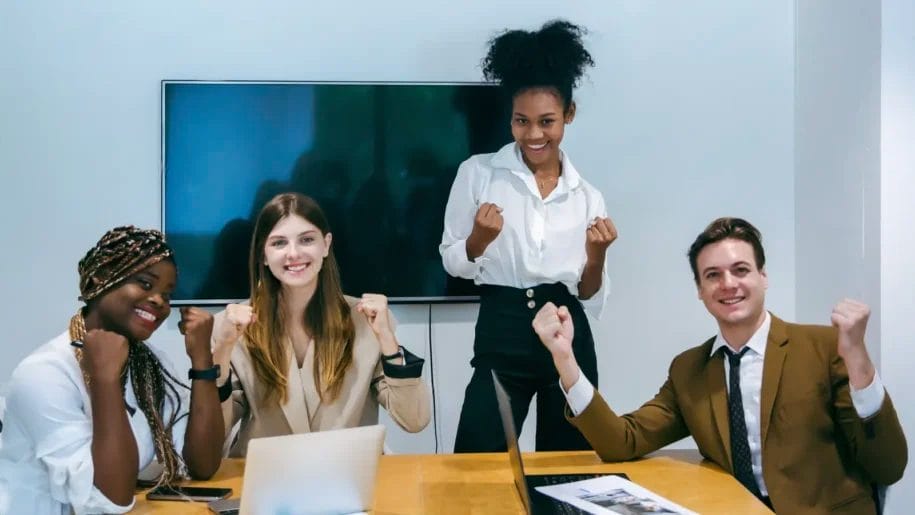 Four people sit and stand around a table with laptops, smiling and raising their fists in a celebratory gesture—a lively scene that could illustrate customer testimonial examples in a modern office meeting room.