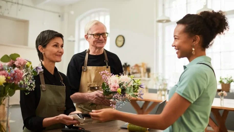 Two florists in aprons stand behind a counter, handing a bouquet to a smiling customer who is paying with a card—an example of excellent customer retention strategies in action. Flowers and the shop’s inviting interior are visible in the background.