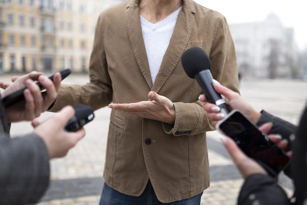 A person in a brown blazer, serving as an example of social proof, gestures with open hands while being interviewed by several reporters holding microphones and smartphones.