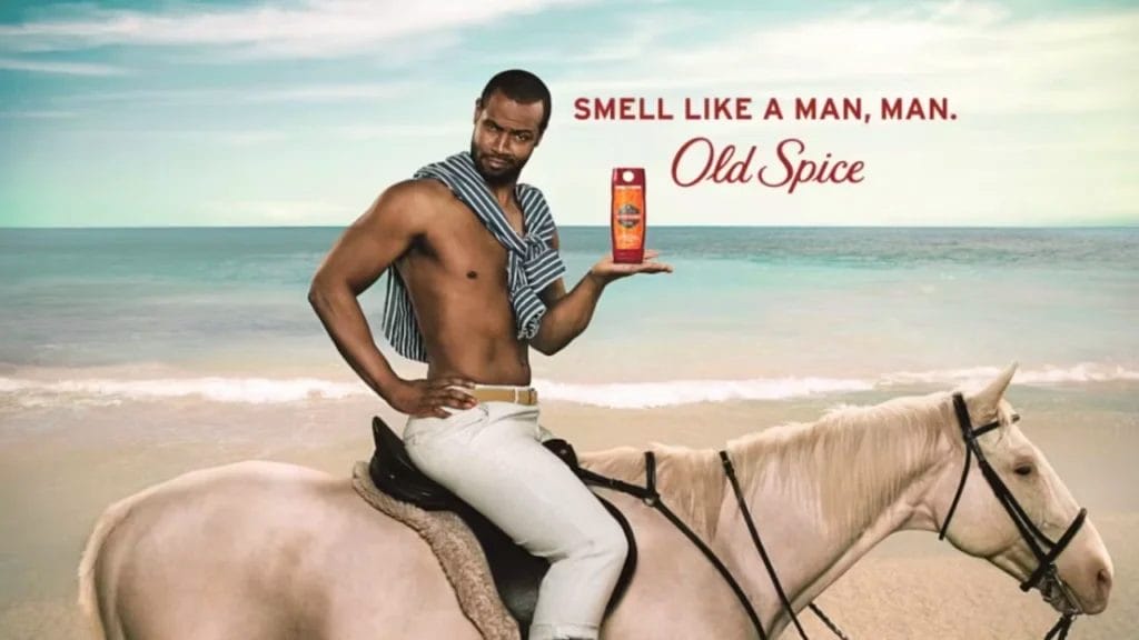 A man sits confidently on a white horse at the beach, clutching Old Spice body wash. Dressed in light pants with a towel draped around his neck, he epitomizes the essence of a viral marketing campaign example. Text reads, "Smell Like a Man, Man. Old Spice.