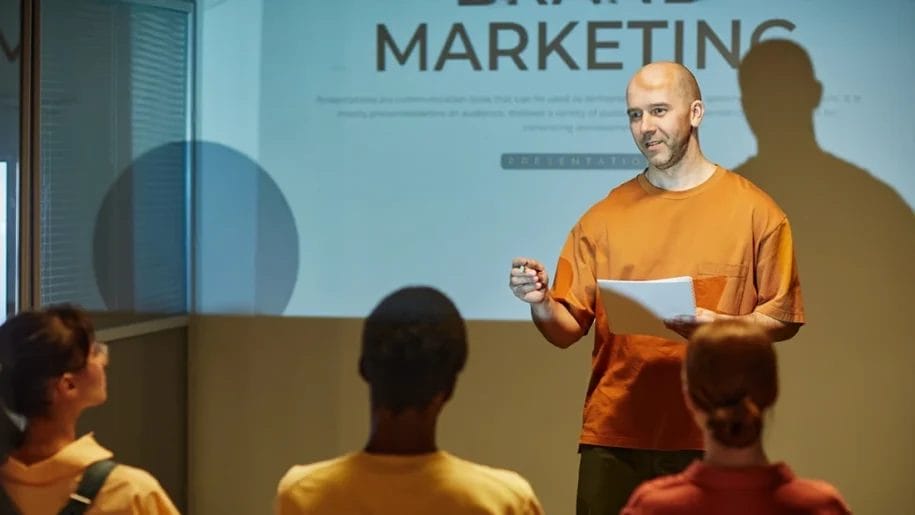 A man stands in front of a projected "Brand Marketing" slide, holding papers and speaking to an audience of three people in a classroom or seminar setting about consumer behavior marketing strategies.