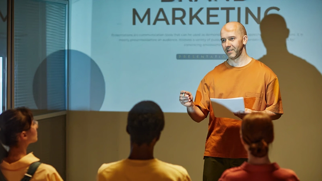 A man stands in front of a projected "Brand Marketing" slide, holding papers and speaking to an audience of three people in a classroom or seminar setting about consumer behavior marketing strategies.