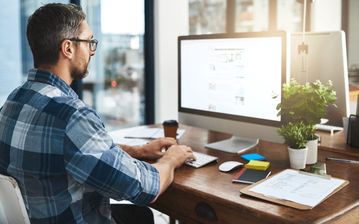 A man wearing glasses and a plaid shirt works on a desktop computer at a wooden desk, surrounded by office supplies, papers, and a potted plant, efficiently handling contact management tasks.