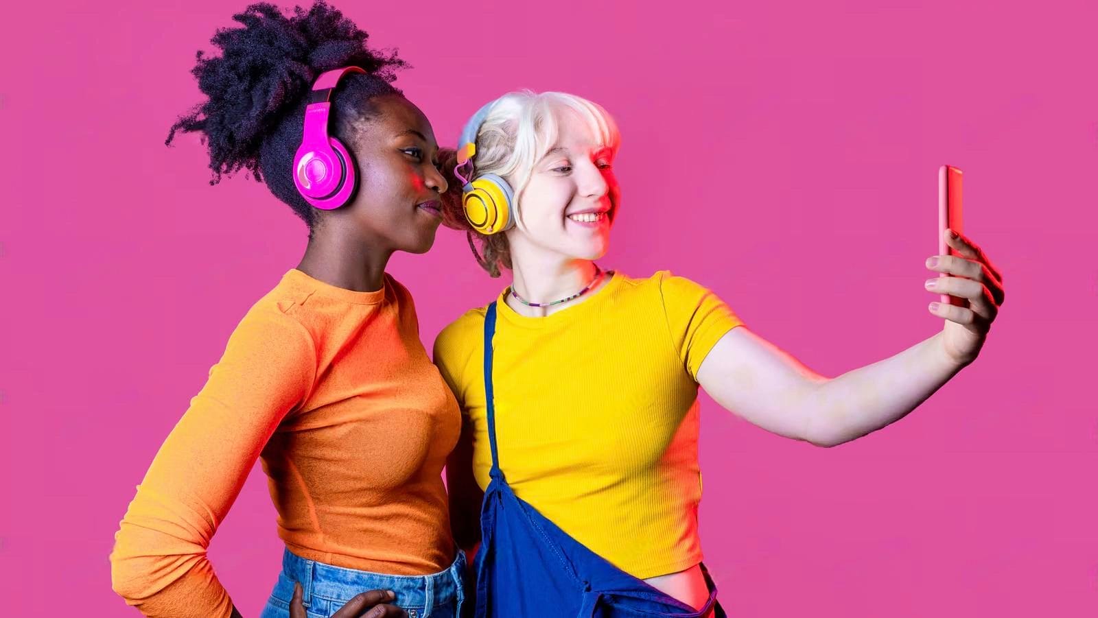 Two young women wearing headphones and colorful shirts take a selfie together against a solid pink background, capturing the perfect moment and showing off their understanding of fomo meaning in text.