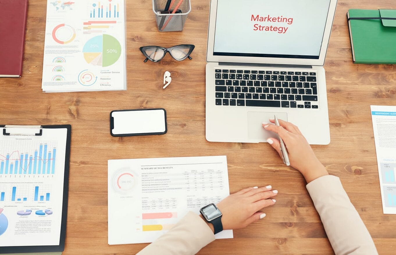 Person working at a desk with marketing charts, a laptop displaying “Marketing Strategy,” documents, and office supplies — analyzing conversion data and reviewing a customer testimonial as a type of social proof.