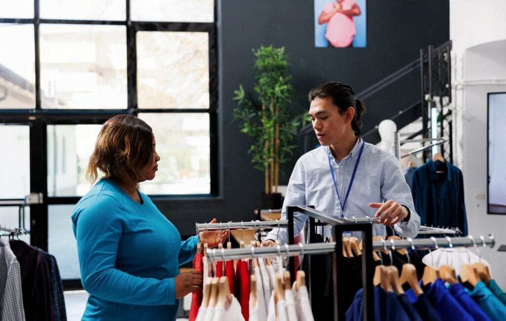 A woman and a store employee stand by clothing racks, discussing garments in a modern retail store that utilizes cutting-edge solutions from leading SaaS companies.