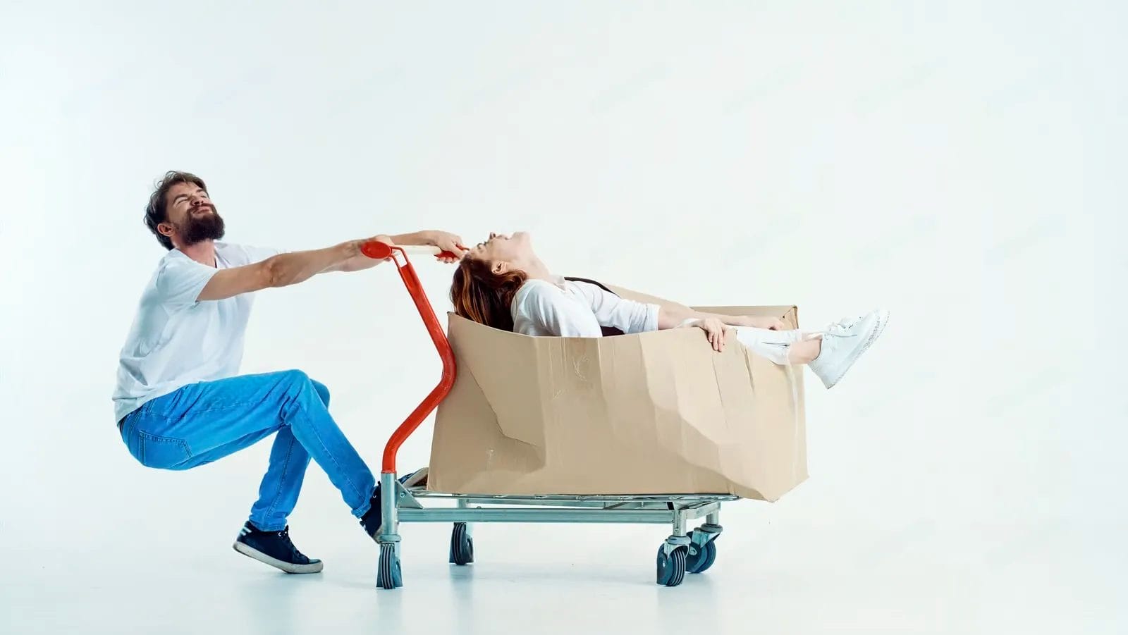 A man pulls a shopping cart with a woman lying inside a large cardboard box; their playful antics in a minimalist studio setting hint at creative ideas on how to reduce cart abandonment.
