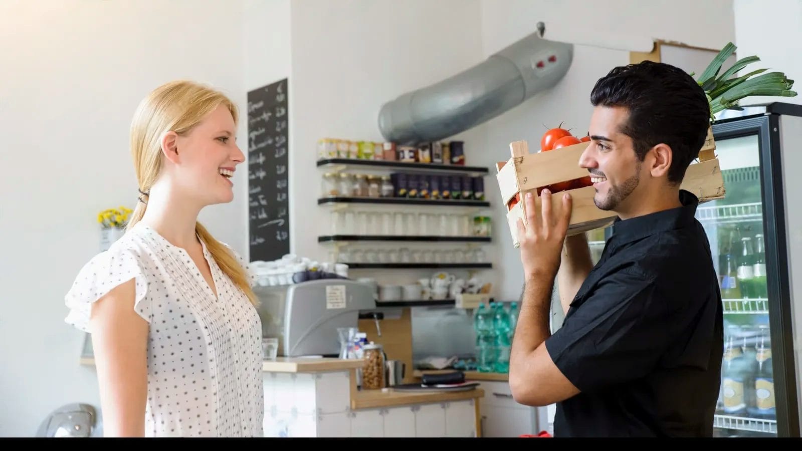 A man holding a crate of vegetables stands in front of a woman in a shop or cafe setting, where shelves with jars, bottles, and a refrigerator are visible—an ideal scene for applying customer feedback analysis methods.