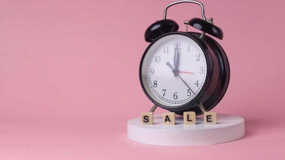A black analog alarm clock on a white pedestal sits next to wooden blocks spelling "SALE," creating urgency in sales against a vibrant pink background.