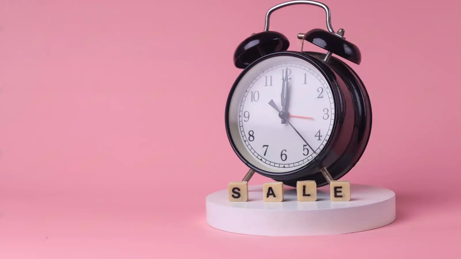 A black analog alarm clock on a white pedestal sits next to wooden blocks spelling "SALE," creating urgency in sales against a vibrant pink background.
