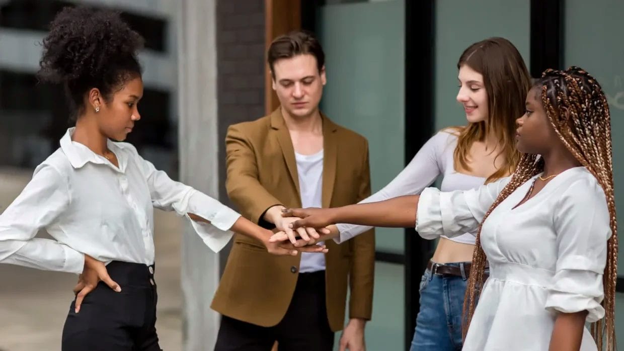 Four people standing outdoors place their hands together in the center, demonstrating teamwork or unity—an informational social influence example of group cooperation.