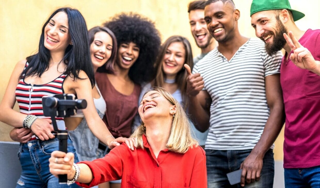 A group of young adults smiling and posing for a selfie taken with a smartphone on a stabilizer, outdoors, capturing the spirit of informational social influence as they follow each other's lead for the perfect shot.
